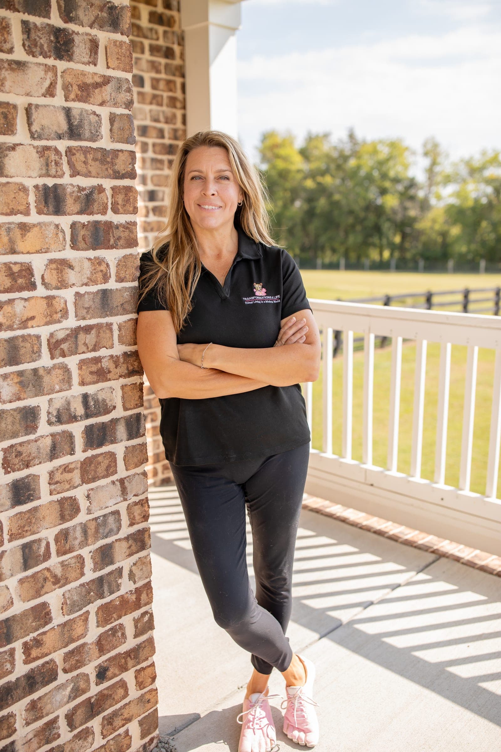 Julie Wilcoxson standing against a brick pillar on a porch, arms crossed, in a black Transformations 4 Life polo and pink barefoot shoes, a fenced field behind her.