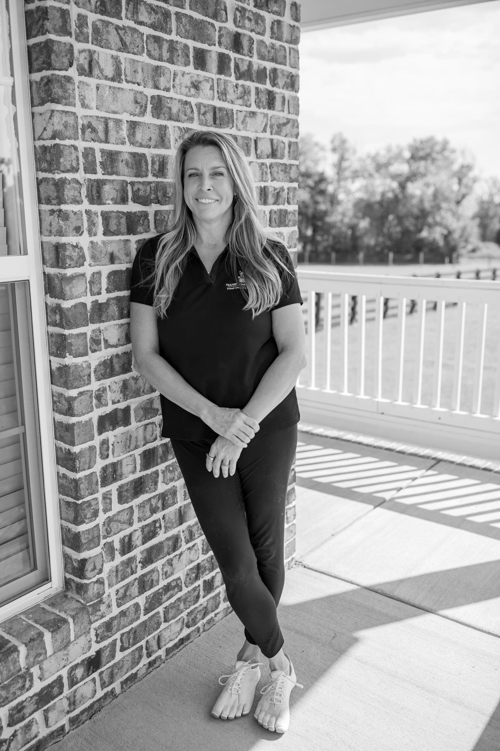 Julie Wilcoxson in black and white, standing against a brick wall on a porch, hands clasped, smiling at the camera.