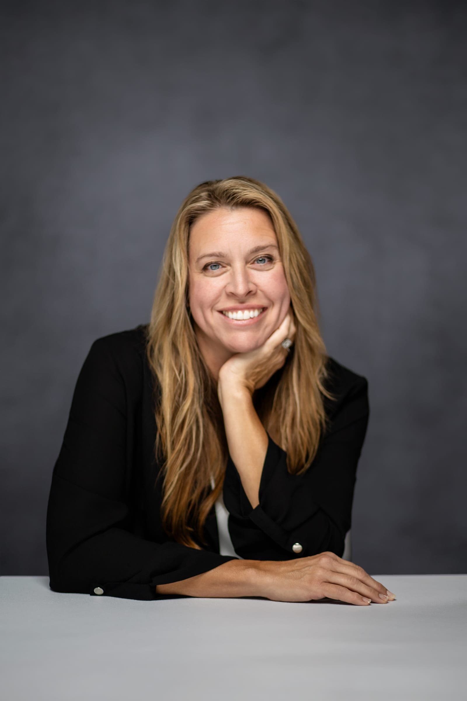 Julie Wilcoxson seated at a pale table in a black blazer, chin resting on her hand, smiling at the camera.