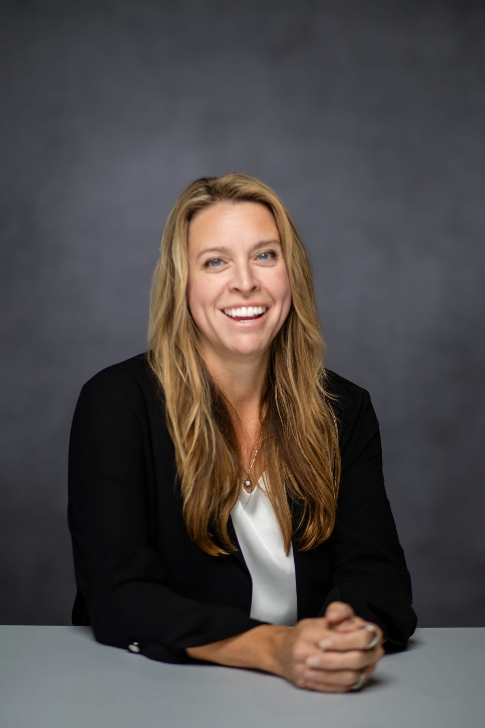 Julie Wilcoxson seated at a table in a black blazer, hands folded, laughing.