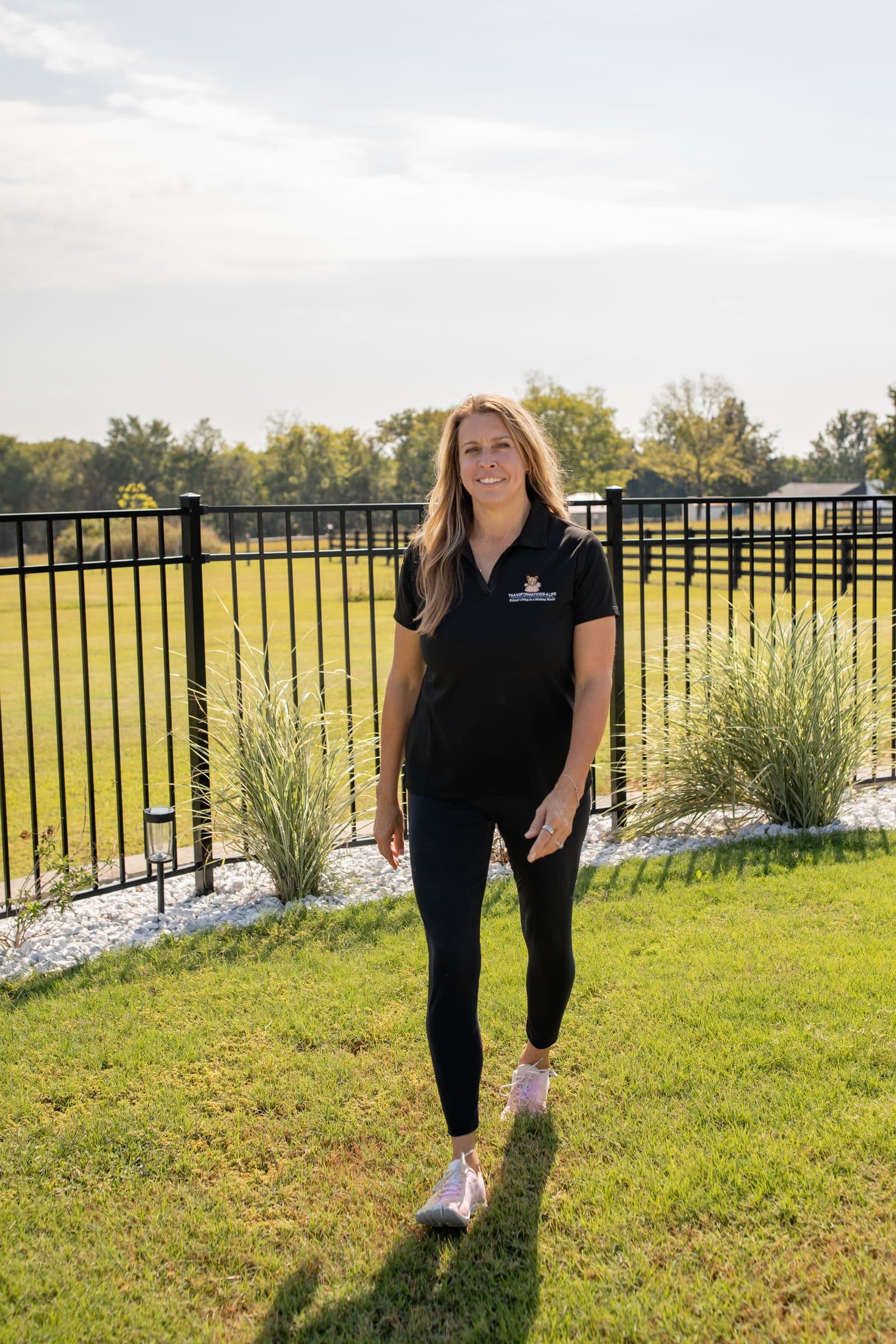 Julie Wilcoxson walking across a lawn beside an iron fence in late afternoon sun, in a black Transformations 4 Life polo and leggings, smiling.