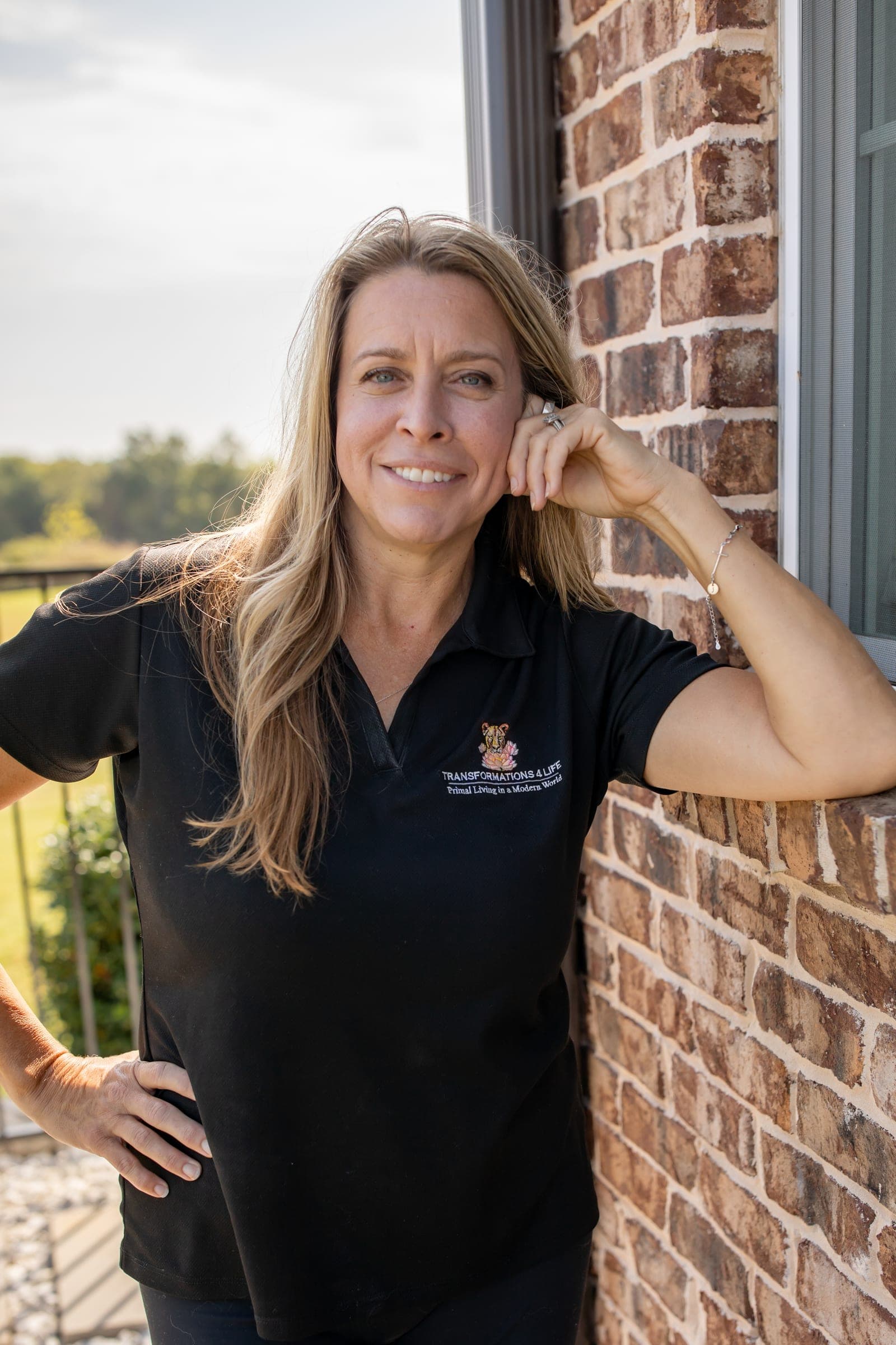 Julie Wilcoxson leaning against a brick wall beside a window, one hand up at her temple, smiling at the camera.