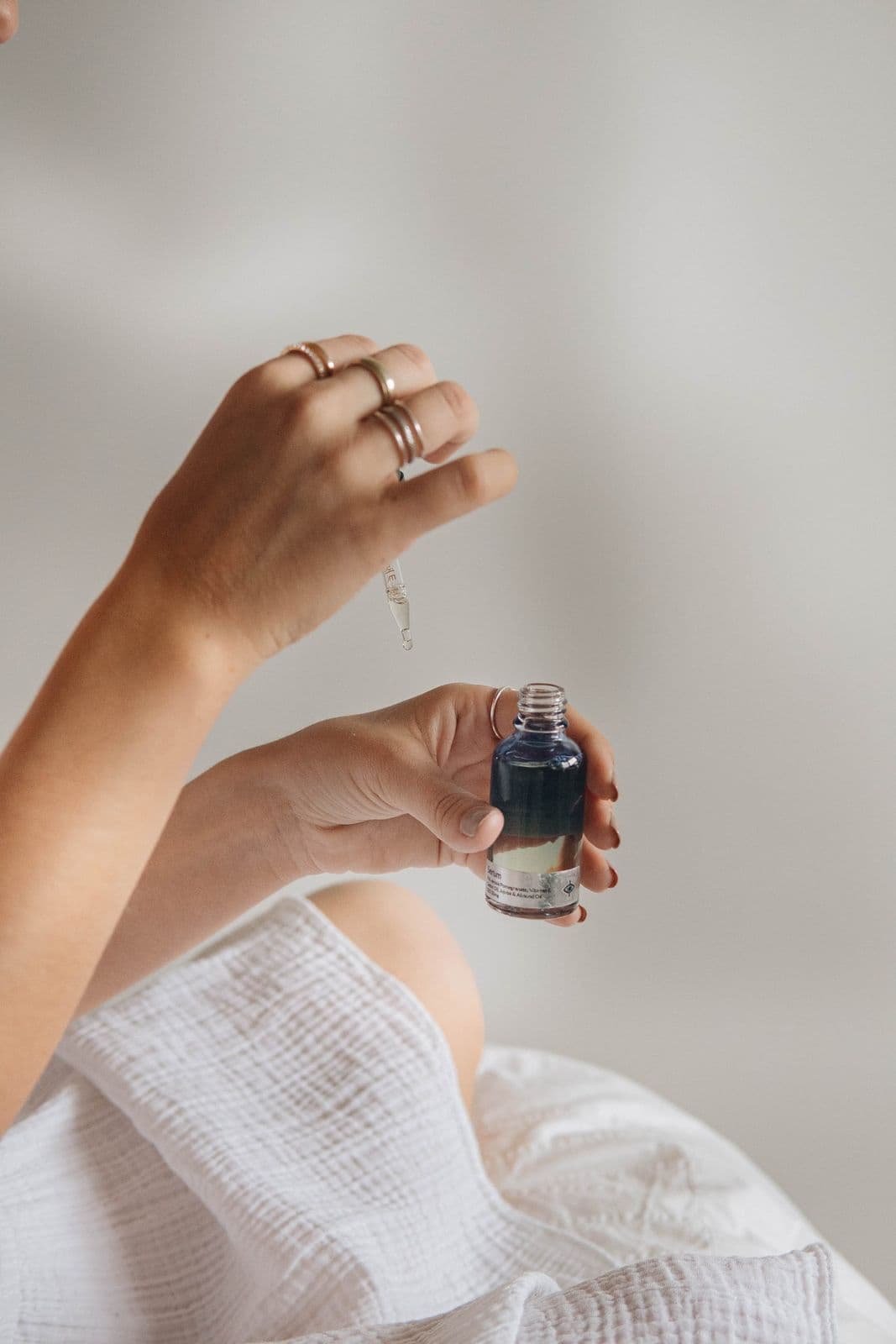 A woman's hands holding a small glass dropper bottle of oil above her lap, white linen draped around her.